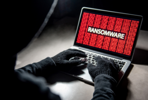 A hacker wearing jacket and gloves typing on a laptop, with a text on the screen showing "RANSOMWARE" and lots of binary characters